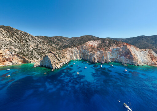 Boats anchored in the turquoise waters of Agios Ioannis Bay on Milos, Greece, surrounded by colorful volcanic cliffs along a popular boat‑tour route.