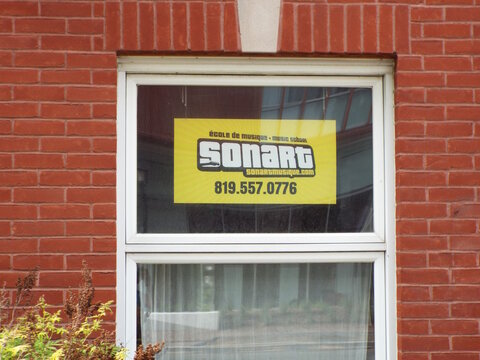 Gatineau,Quebec, Canada &ndash; June 4, 2026: A vibrant yellow sign for "Ecole de Musique Sonart" featuring a telephone number hangs inside a window of red brick on rue Principale.