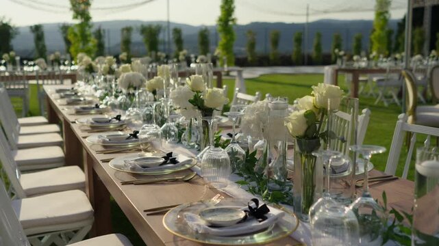 Long wedding table lined with white roses gold place settings and glassware on a green lawn