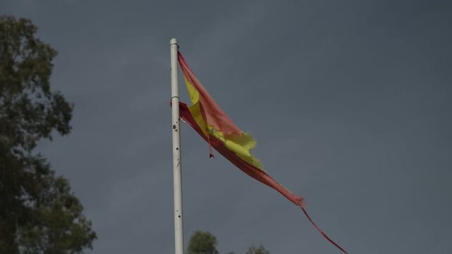 Old faded and tattered Spanish flag frayed on a white flagpole against an overcast grey sky