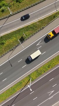 Vertical screen. Aerial perspective shows a large truck and a smaller car moving along a multi-lane highway, with green grass and trees lining the road under bright sunlight on a clear day