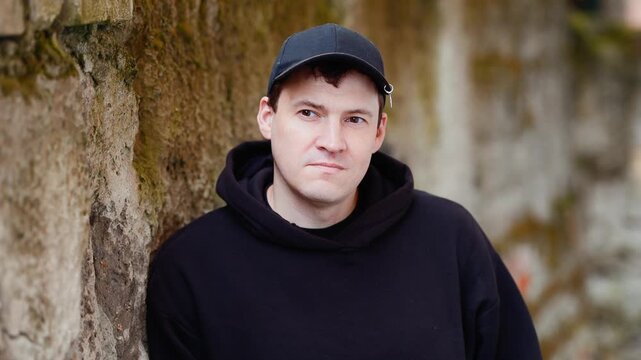 Man in black hoodie and cap leaning against rustic stone wall. Casual portrait. Urban street style outdoor session.