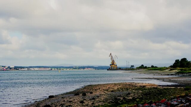 Coastal view of Firth of Clyde in Greenock Scotland. Calm water, rocky shoreline and industrial crane stand beneath cloudy sky along the estuary coastline