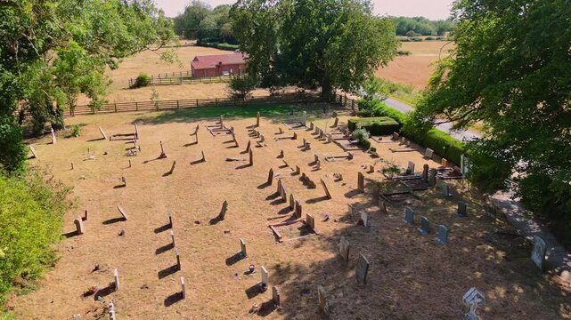Aerial view of an old cemetery surrounded by fields and trees. Weathered headstones lie in quiet rows under soft sunlight. Lone car passes on a nearby road, adding subtle motion
