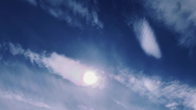 A view of a clear, bright blue sky with intense sunlight at its center. White clouds drift slowly, creating a dramatic, serene, and natural atmosphere.