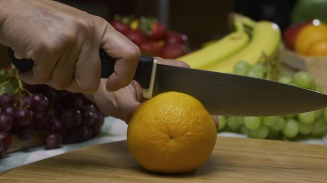 Slow motion shot of knife cutting through delicious, juicy orange on cutting board near other fruit - closeup