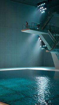 Indoor high diving competition with illuminated pool water, athlete on diving board and splash reflection aerial view