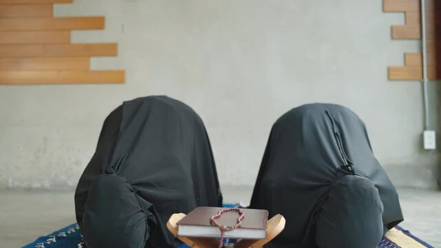 Two Muslim women performing sujood during Islamic prayer with the Quran placed on a wooden stand and prayer beads in front. Worship, devotion, humility, and spiritual faith concept.
