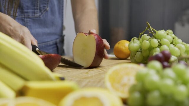 Female cook slicing through apple with sharp knife on cutting board with surrounding fruit - closeup slow motion shot