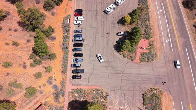Aerial drone footage of Utah Scenic Byway 12 capturing a sunlit parking area with cars, RVs, and recreational vehicles nestled beside orange desert terrain with scattered green juniper trees.