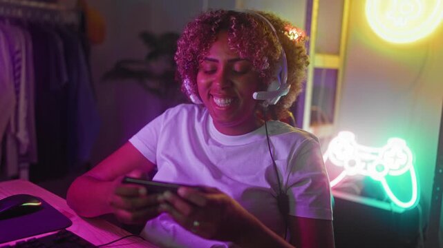 Woman wearing headset smiling and holding smartphone while gaming at desk with keyboard and neon controller sign in studio; joyful connection.