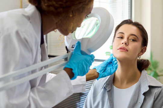 Dermatologist examination with patient in clinic consultation. Gloved doctor uses magnifying lamp to assess skin, discuss concerns, and recommend treatments. Concept shows expert skin care.