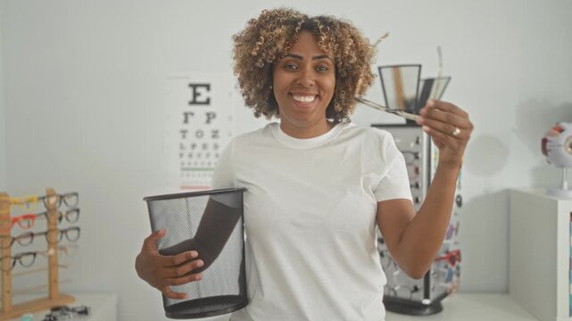 Woman holding eyeglasses and mesh waste bin, smiling and giving thumbs up while sorting frames in an optical studio; joy.