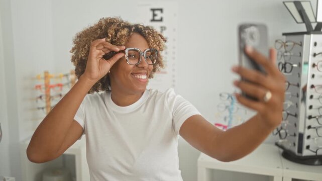 Woman adjusting glasses while holding smartphone for a selfie in an optical store inside a building; self expression joy.