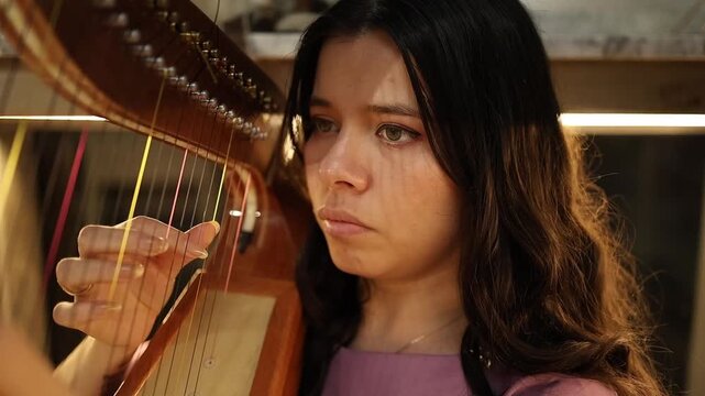 A focused young woman plays the harp with delicate precision in a warmly lit room, her expressive face showing concentration. The close-up shot highlights her hands and strings in rich detail.