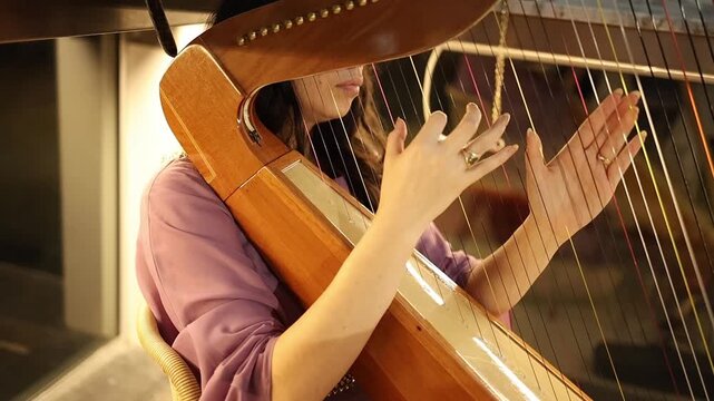 Elegant close-up shot of a woman in a lavender dress skillfully playing a wooden harp indoors. Warm, ambient lighting highlights the movement of her hands and the harp's shimmering strings.