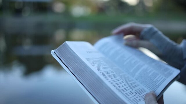Close-up shot of hands turning pages in an open book beside a tranquil lake, natural daylight, shallow depth of field, relaxed atmosphere, outdoor reading, serene reflective mood.
