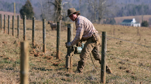 Man farmworker using post hole digger and tools to erect fence posts with barbed wire in farmland countryside