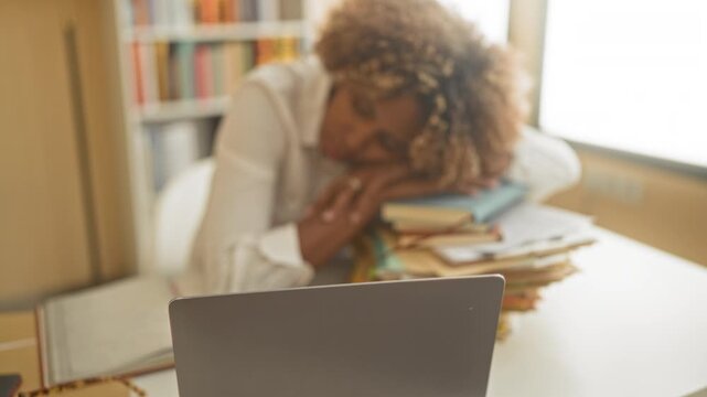 Young african american woman with head resting on a stack of books and hands folded over papers at a laptop in a building study; exhaustion study burnout.