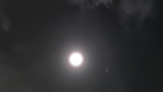 A luminous full moon glows brightly in the night sky, encircled by a faint halo of light. Wisps of scattered clouds drift across the upper frame, softly illuminated by the moonlight.