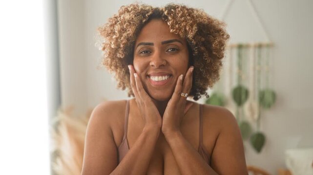 Woman smiling with hands on cheeks in studio setting showing bra straps and bare shoulders, warm natural light and houseplants in scene; self care joy.