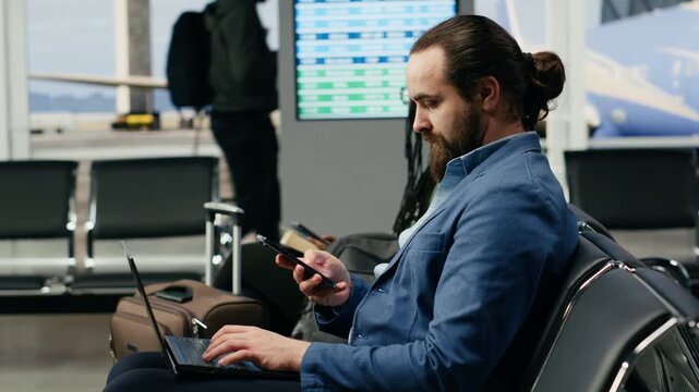 A businessman uses a laptop while waiting for a smartphone message at the airport, checking notifications and completing computer tasks before departure on an international business flight.