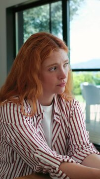 Worried and stressed redhead young woman at home looking outside at the window thinking about personal problems