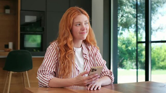 Beautiful young woman with long red hair sitting at a table in her modern kitchen, thoughtfully scrolling through her phone, her expression shifting from serious to a gentle, happy smile