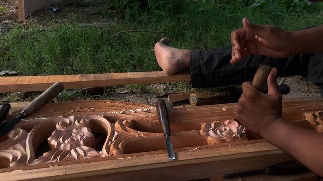 Closeup of craftsman carving intricate floral pattern into wooden panel with chisel and mallet, shaping decorative relief by hand during woodworking process.