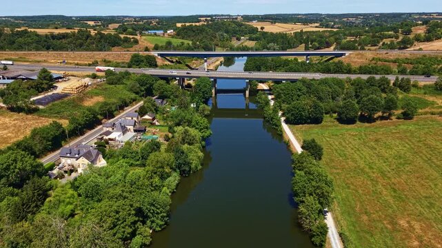 Forward drone flight above the Mayenne River approaching the L'Armoricaine highway bridge with moving traffic, surrounded by rural landscape, trees and farmland in Chang&eacute;, France.