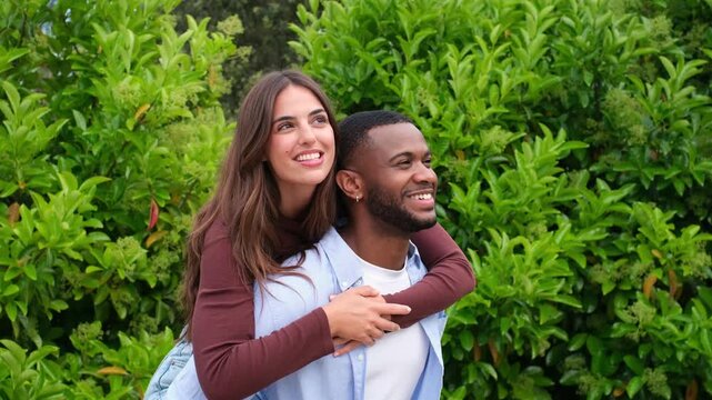 Young multicultural couple enjoying a romantic moment outdoors as the man gives his girlfriend a piggyback ride while she points at something, both smiling and having fun together in a park