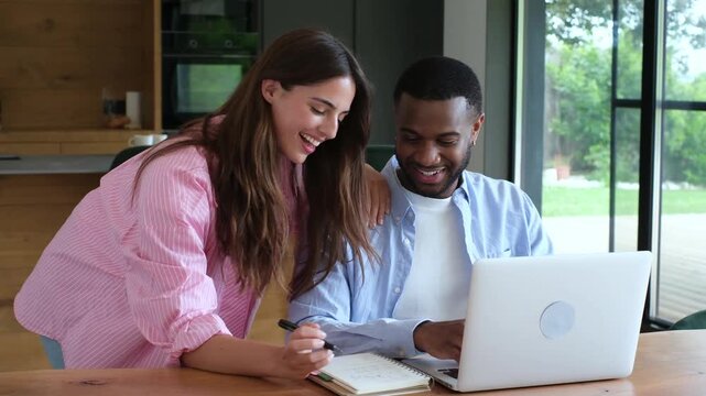A young, happy couple collaborates at home, smiling and taking notes while working on a laptop. They share a moment of productivity and teamwork in their modern living space.