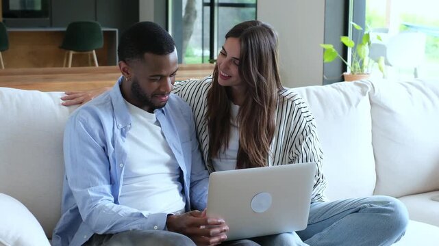 Happy young mixed-race couple sitting together on the couch at home, watching funny videos on a laptop, browsing the internet, making online purchases, or planning a vacation trip