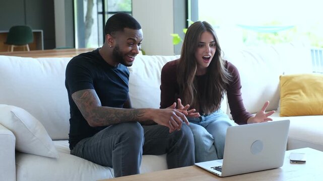 Young overjoyed multiethnic man and woman sitting on the couch, looking at a laptop screen, celebrating a win with a high five, expressing happiness, and enjoying their shared success at home