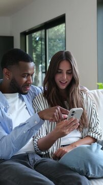 A happy and diverse couple relaxes on their couch at home, enjoying content together on a mobile phone. They smile and laugh, sharing a joyful moment.