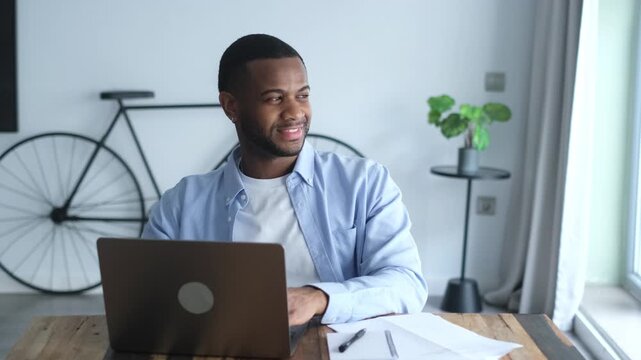 Young handsome african american man sitting at his desk, typing on a laptop and thoughtfully looking out the window while working on a project from his modern, stylish home office