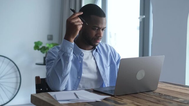 A focused young Black man works diligently at a wooden table in his home office. He thoughtfully uses his laptop and takes notes, demonstrating dedication to his task.