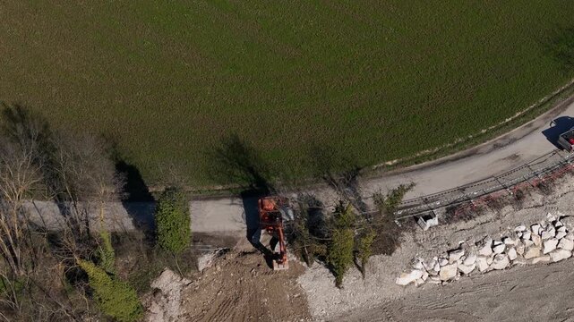 Hydraulic excavator performs riverbank earthworks beside a gravel access road and riprap embankment, supporting bank stabilization, flood protection and civil engineering infrastructure, top-down