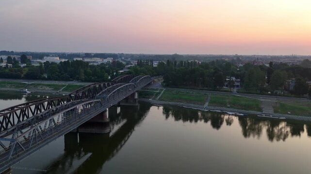 Ponte di Ferro railway bridge spans the Po River between Cremona, Lombardy and Castelvetro Piacentino, Emilia-Romagna, Italy, beneath a colorful sunrise sky with calm reflections drone pullback