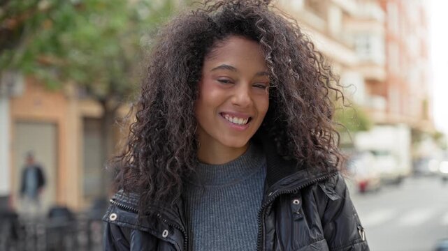 Teenage woman smiling showing teeth on a street, wearing jacket and sweater with curly hair framing her face; joy.