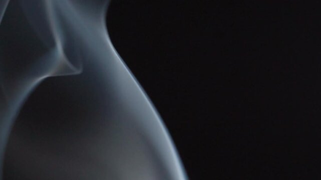 Closeup of delicate incense smoke swirling gracefully against black background. Soft flowing patterns creating elegant abstract movement with smooth curves and fine wisps