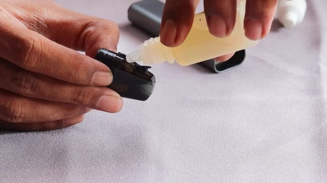 Close-up of an Asian man's hands refilling vape pod with e liquid from dropper bottle, detailed view of pod maintenance and modern electronic cigarette use.