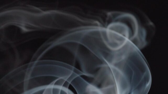 Abstract white incense smoke drifts across a black background, forming soft flowing patterns. Macro closeup captures delicate curves, gentle movement and atmospheric wisps in the air