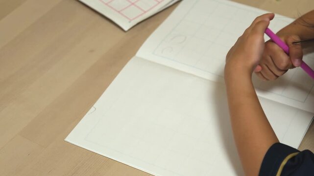 Close-up of a child's hands writing in a notebook with a pencil at a desk