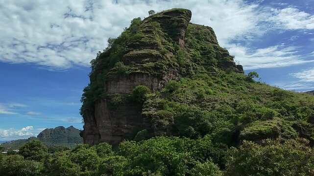 Aerial drone shot of Cerro Hotcakes rock formation rising above lush tropical vegetation under dramatic clouds during daytime in Morelos, Mexico.