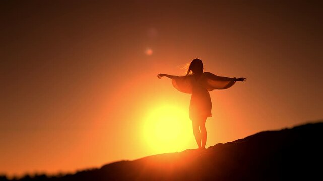 Graceful silhouette of young woman spinning with outstretched arms on sandy hill during golden sunset. Artistic lifestyle scene symbolizing freedom and joy.