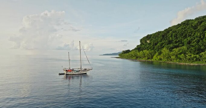 Sailboat yacht peacefully anchoring in calm blue ocean waters offshore lush tropical jungle coastline of Tegua island, offering a serene and secluded travel experience to Vanuatu. Aerial drone flight