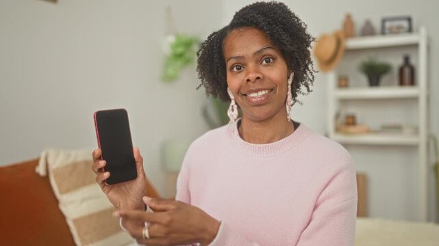 Woman holding smartphone and pointing to its blank screen while seated on a bed in a bedroom, smiling and presenting the device; friendly demo.