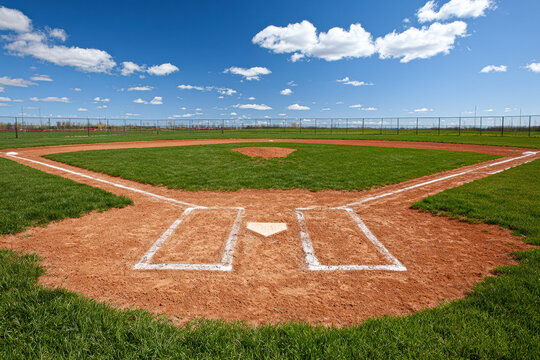 Empty baseball field under a blue sky with scattered clouds, showing home plate and infield dirt with green grass beyond.