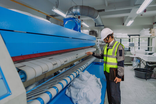 Professional Asian industrial worker in safety gear holding a tablet while inspecting a heavy-duty flatwork ironer machine at a modern commercial laundry factory for hotel service.
